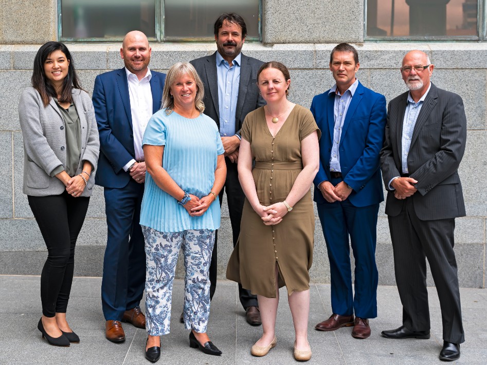 A team of people in office-wear stand together in front of a grey building wall.