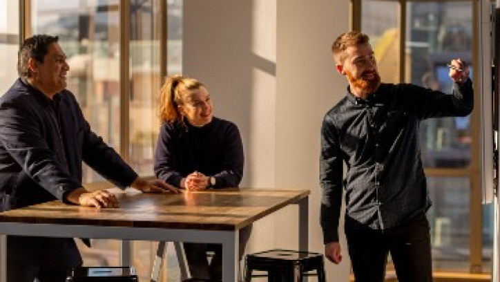 A bearded person is pointing to a presentation while two others watch while smiling.